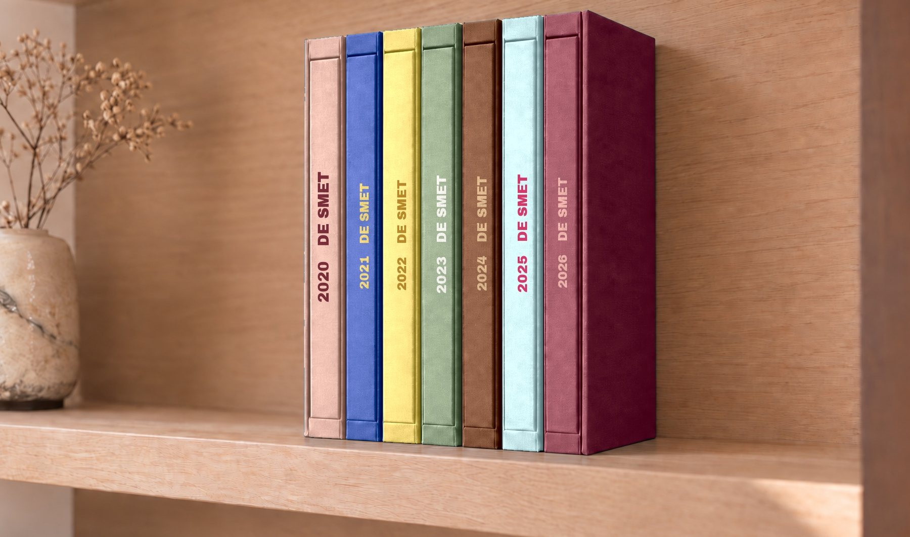 Zeven familie-albums naast elkaar op een eiken plank, elk jaar in zijn eigen kleur, met jaartal en naam op de rug